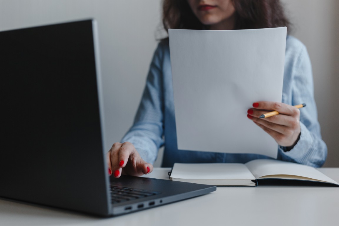 Closeup of woman's hand with red nails holding documents and sitting in front of a laptop wearing blue shirt. Business, freelance, self employment. Distance job and online work concept.